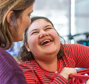 A young woman standing in adaptive equipment and holding a red basket shares a moment of laughter with her therapist.
