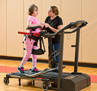 A young girl gait trains on a treadmill in a gym with the assistance of a therapist.