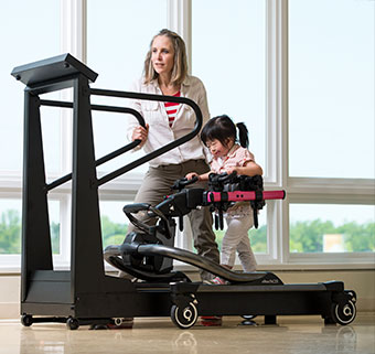 A young girl uses the treadmill base and pink Rifton Pacer to gait train over a treadmill while assisted by her therapist.