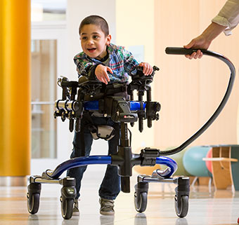 A child with sci in a blue dynamic gait trainer smiles at his caregiver as he walks through the school hallway.