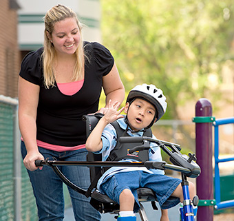 A caregiver steers a child in a blue Rifton Adaptive Tricycle.