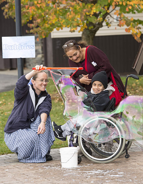 A young woman blows bubbles for a child in a wheelchair while his mother looks on.