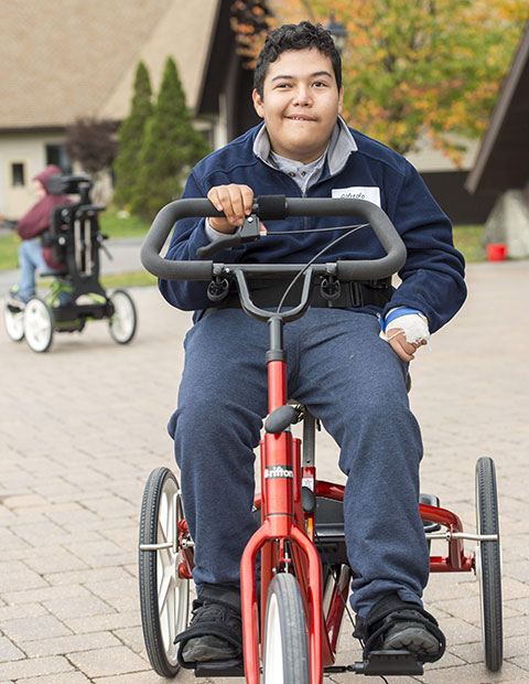 A young man rides on a red adaptive tricycle participating in rifton's birthday tricycle raffle.