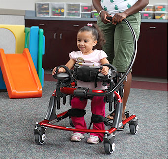 A young girl with cerebral palsy walks in a red Pacer in therapy while her therapist stands behind holding the guide bar.
