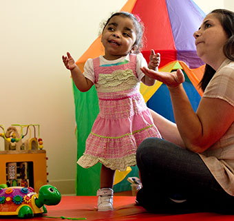 A young girl practices walking on a red mat while her therapist looks engages her in motor learning strategy. 