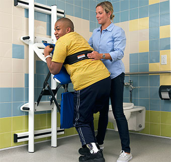 A therapists assists a young man in a sit to stand transfer for toileting at the Support Station.