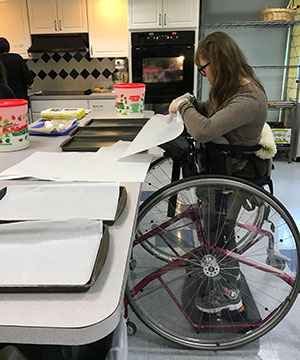 A young woman uses adaptive equipment to stand at the kitchen counter during a life skills class where she is placing baking sheets on trays.