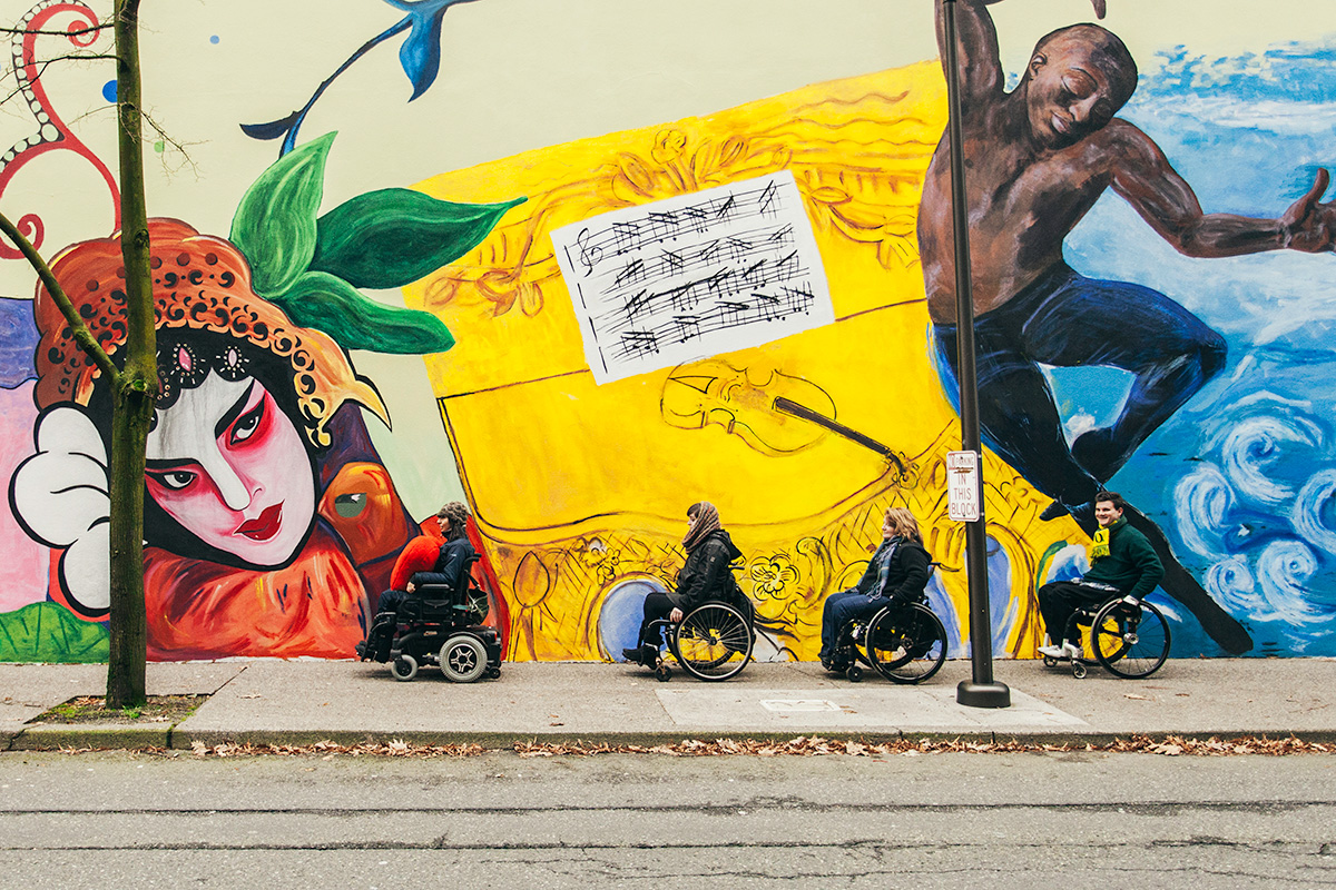 Four wheelchair users, the lead of which has a heart-shaped pillow in her lap, roll in front of a brightly colored mural 