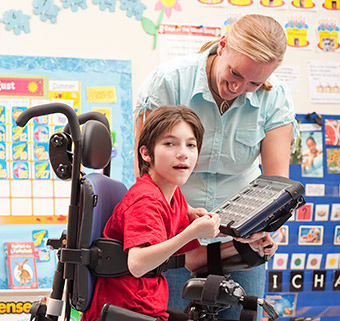 A young boy sits in a Rifton Activity Chair and uses an assistive device to talk to his caregiver.