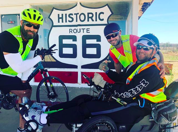 Three men in helmets, one on an electric smile in front of a Route 66 sign during the cross-country ride for paralysis 