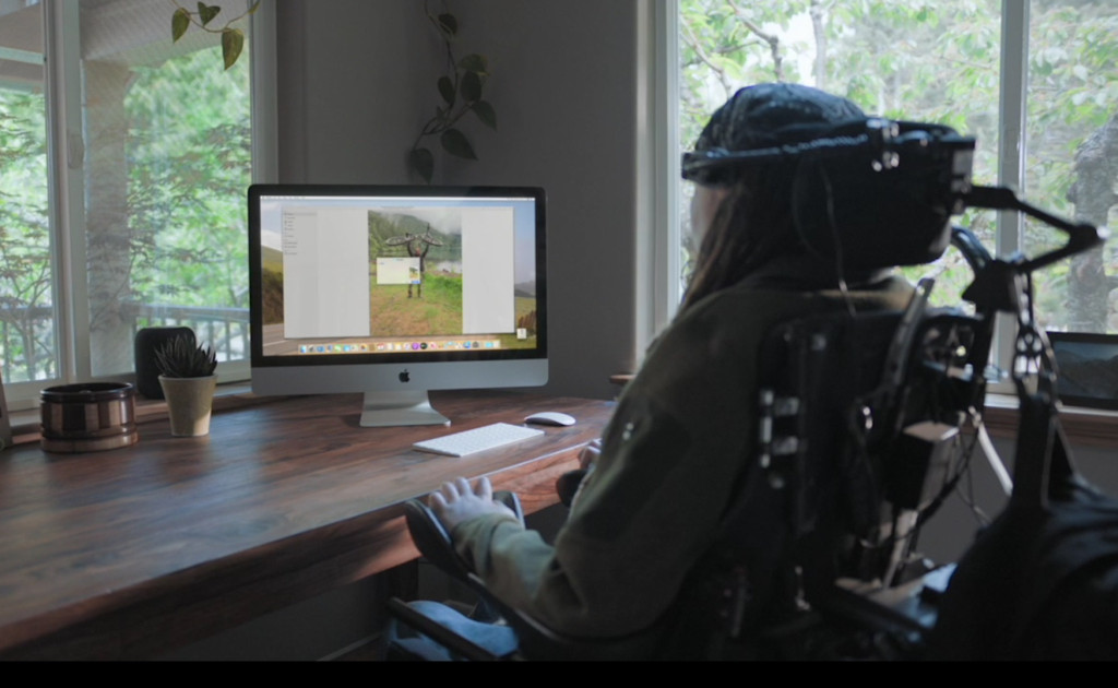 Man sitting in power wheelchair shown using Voice Control, an accessibility feature, on his Apple desktop computer