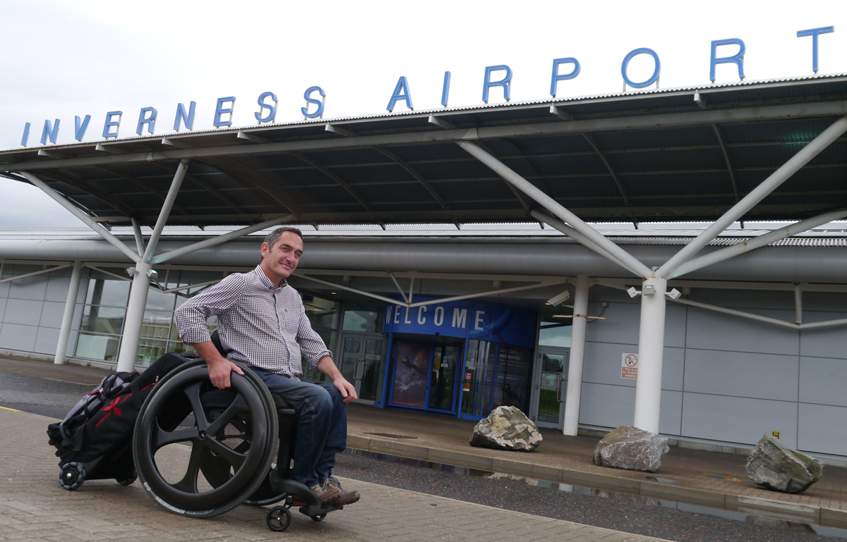 A man in a wheelchair in front of airport with roller bag on his chair, airlines now committing to improving disabled travel