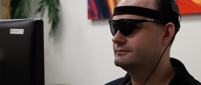 a person with acquired blindness is seen wearing sunglasses with camera attached to them. He is sitting in front of a computer.