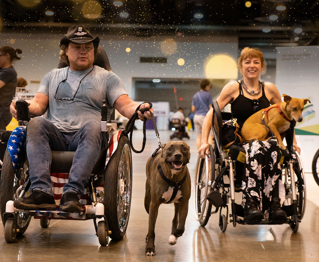 Two wheelchair users at the Abilities Expo, male wearing a cowboy hat with a dog on a leash running to his side, and a female with a dog on her lap, pushing a manual wheelchair.