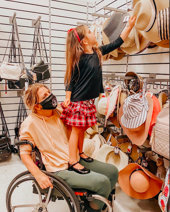 photo shows woman in wheelchair wearing facemask, shopping with daughter standing on her lap to reach a hat on high rack