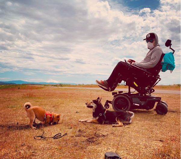Image of man in power wheelchair wearing mask with two dogs in a field, submitted to New Mobility photo contest