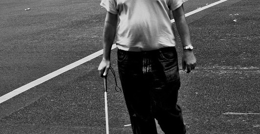 A blind man walking on the road with a white cane in his right hand